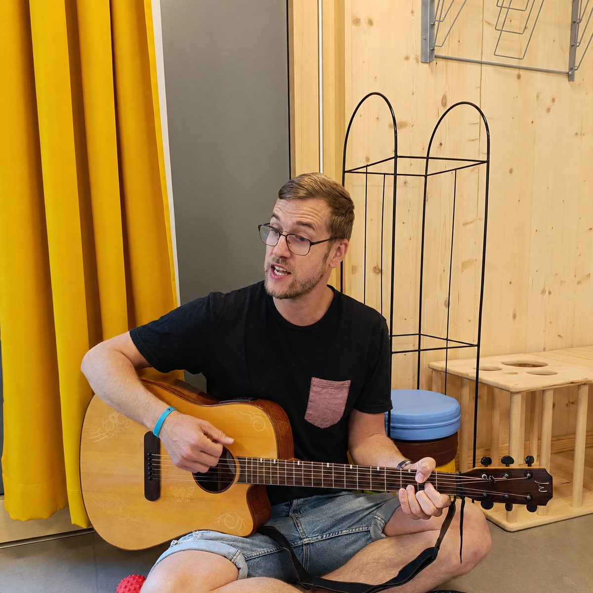 Heilerziehungspfleger Alex sitzt mit einer Gitarre in einem Gruppenraum einer Kinderkrippe und singt
