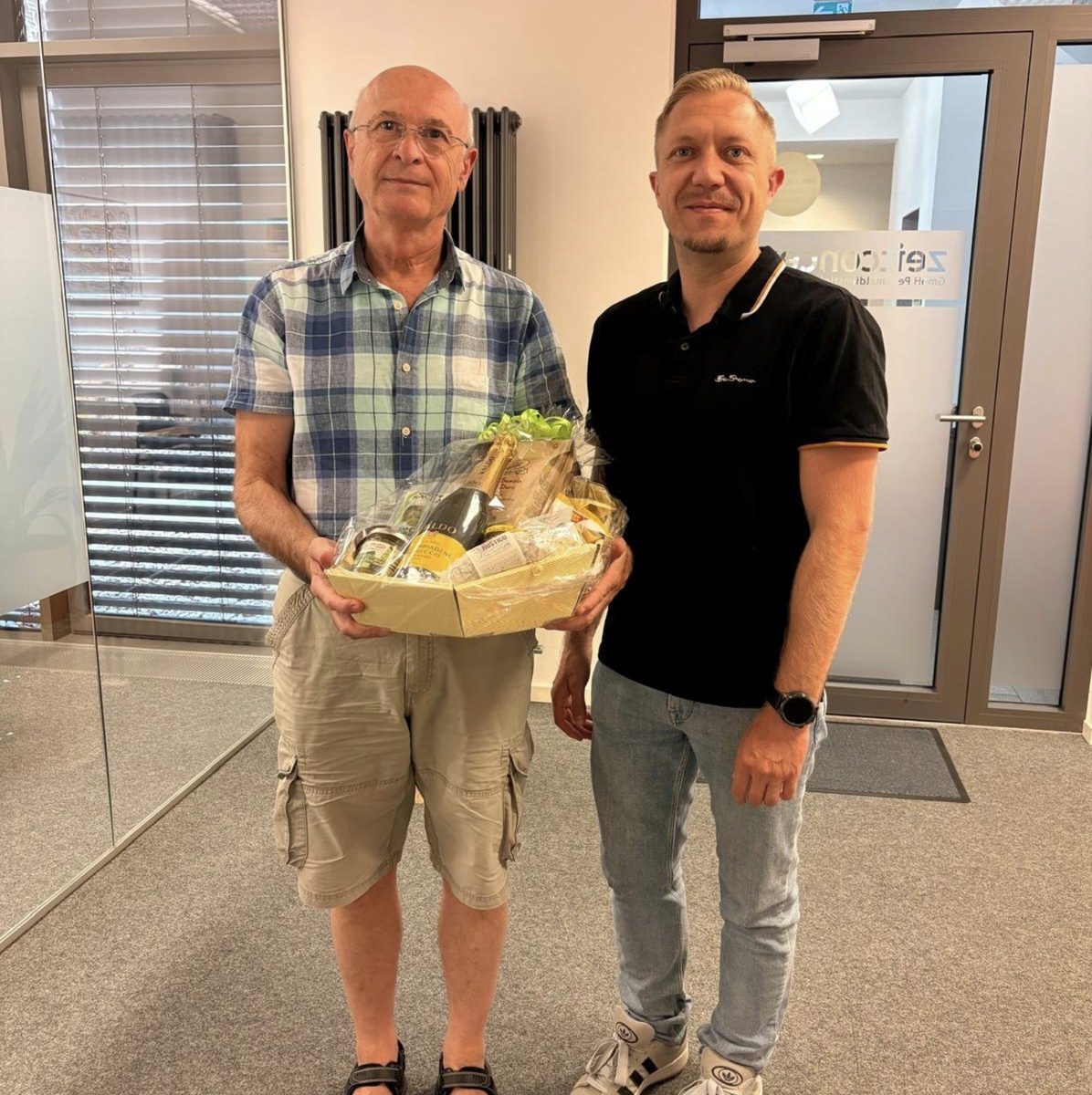 Herr Schimpf mit einem Präsentkorb, daneben ein Kollege, bei der Verabschiedung in den Ruhestand in der zeitconcept-Geschäftsstelle Regensburg
