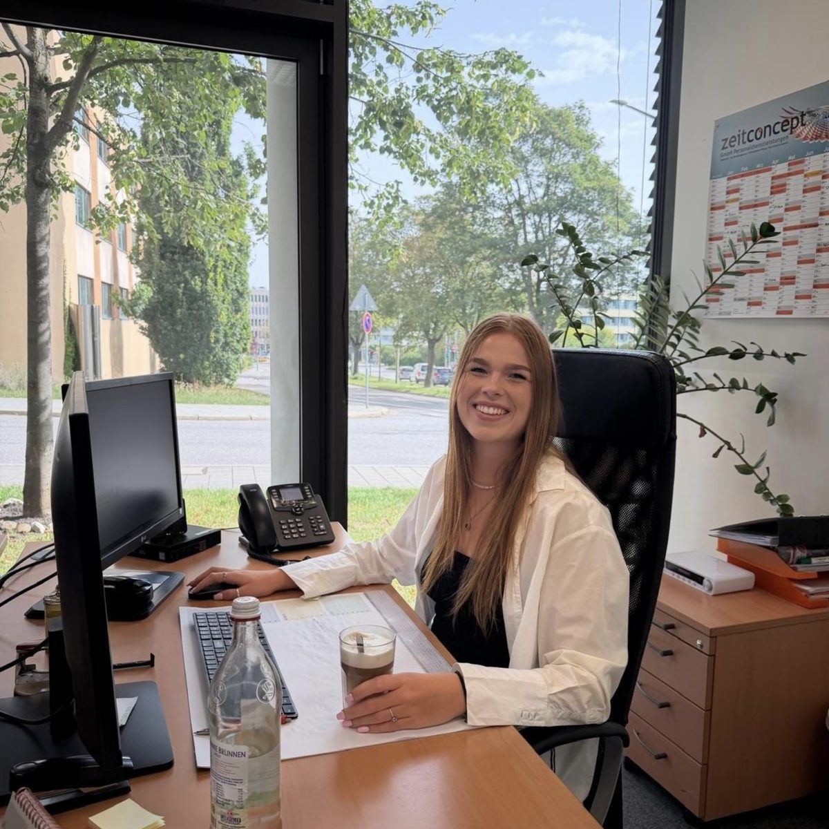 Verena sitzt lächelnd an ihrem Schreibtisch in der zeitconcept-Geschäftsstelle Regensburg; vor ihr Bildschirm, Tastatur, Telefon und ein Milchkaffee, am Fenster der Blick ins Grüne, im Hintergrund ein zeitconcept-Plakat